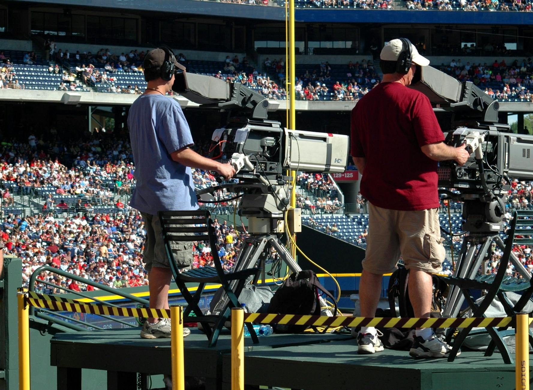 Camera operators capturing a live sports event at a crowded outdoor stadium filled with spectators.